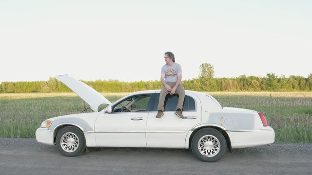 Man sitting on top of his car after it broke down in the middle of nowhere. Looking desperate.