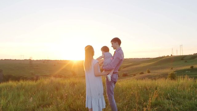 Young happy family with baby. They go to the picturesque green field at sunset.