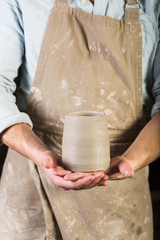 pottery, stoneware, ceramics art concept - young man in a dirty apron stand at workshop, master hands holds cup of unfired clay, male examines product before further processing, front view, vertical