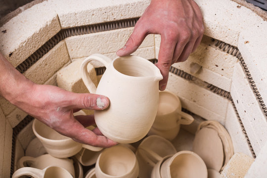 pottery, workshop, ceramics art concept - top view of electric oven for roasting of unbaked clay products, male hands putting the jug in the kiln, some unfinished cups and utensils