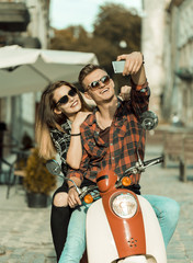 Obraz na płótnie Canvas Young attractive couple of teenagers wears sunglasses on retro motorbike makes selfie, sunny street, urban hipster concept