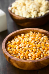 Yellow raw popcorn kernels in wooden bowl with salted popped popcorn in the back, photographed with natural light (Selective Focus, Focus in the middle of the kernels)