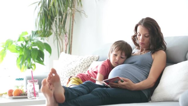 Young pregnant woman, reading a book at home to her boy, eating fruits, hugging and laughing