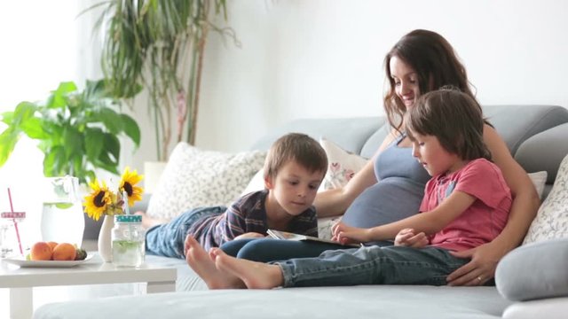 Young pregnant woman, reading a book at home to her two boys, eating fruits, hugging and laughing