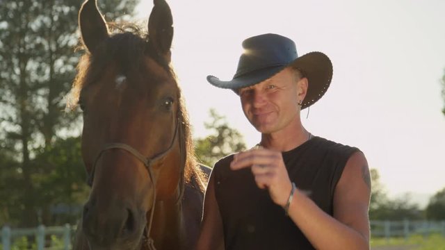 SLOW MOTION, CLOSE UP, DOF: Happy elder rider wearing cowboy hat, greeting, touching his hand to the brim. Horse shaking his head on sunny spring day. Portrait of a senior man and a stallion at sunset