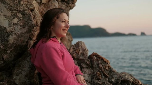 close shot of woman face on shore of lake, looking on landscape. Attractive female with brown hair sitting under rocks, mountains. Closeup shooting nice lady in pink sweater, natural makeup walks on