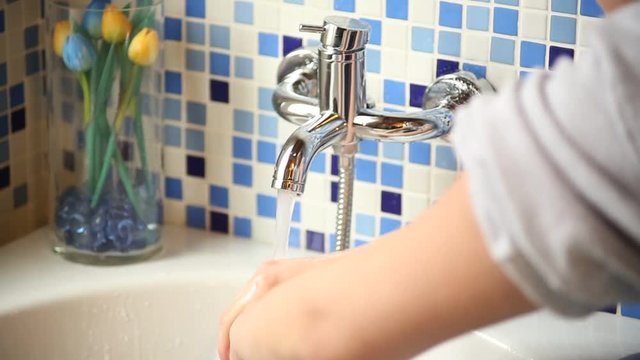 Young woman is cleaning his hands with water at the tap of a bathtub. The wall is gres tiled with blue and white tiles.