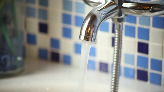 Water tap of a bathtub opening and closing, comes out hot water and steam. The wall is gres tiled with blue and white tiles.
