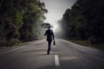 Back view of businessman with umbrella and suitcase walking on road