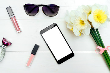 Top view on white wooden background with lip gloss, perfume, sunglasses and phone with white screen around bouquet of narcissus, copy space