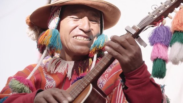 Native quechua man using a colorful handcrafted chullo and a highlander hat, singing with his guitar on the alleys of Cusco	