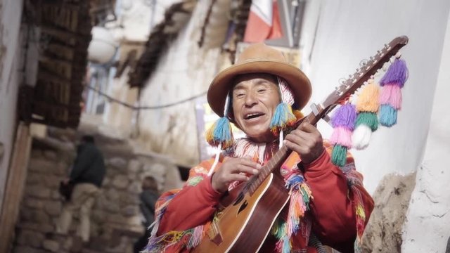 Native quechua man using a colorful handcrafted chullo and a highlander hat, singing with his guitar on the alleys of Cusco