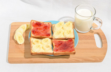 Square toast with jam and banana on a blue plate on a wooden board and a mug of milk