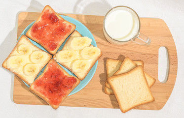 Square toast with jam and banana on a blue plate on a wooden board and a mug of milk