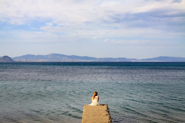 Alone woman in white casual outfit with long hair sitting at beachfront on a small concrete pier.