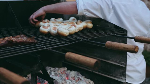 Hand held shot with soft focus of big industrial barbecue tray, roasting beef and burger buns on hot coal, waiter or chef preparing ingridients for serving in restaurant or food truck joint