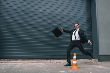Stylish businessman in eyeglasses holding briefcase and running near traffic cone - Powered by Adobe