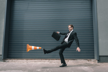 Excited businessman in eyeglasses holding briefcase and kicking traffic cone