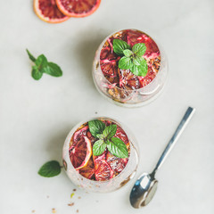 Healthy breakfast. Greek yogurt, granola, blood orange layered parfait in glasses with mint leaves over light grey marble background, top view, square crop. Clean eating, detox, dieting food concept