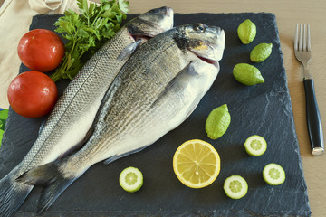 Two ready to cook raw fish with vegetables on stone slate board.