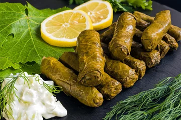 Foto op Plexiglas Delicious stuffed grape leaves (the traditional dolma of the mediterranean cuisine) on black dish with leaves, lemon slices, dill and tzatziki sauce. © dinosmichail