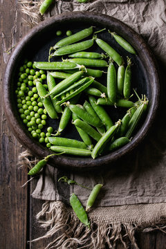 Young organic green pea pods and peas in terracotta tray over old dark wooden planks with sackcloth textile background. Top view with space. Harvest, healthy eating.