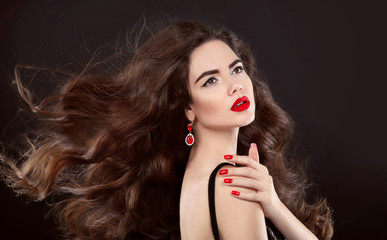Fototapeta premium Beauty hair. Red manicure. Brunette girl with makeup and long shiny wavy hair. Fashion earring jewelry. Beautiful model portrait with curly hairstyle, manicured nails over dark background.