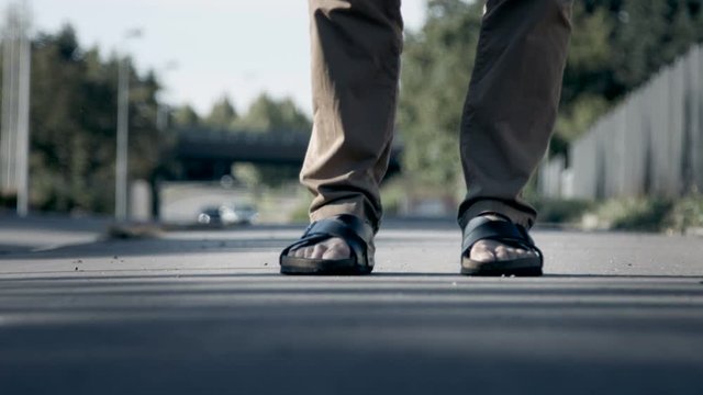 A man wearing a pair of sandals taps his feet and makes a stomp. Filmed from ground level and with a background of traffic in shallow focus.