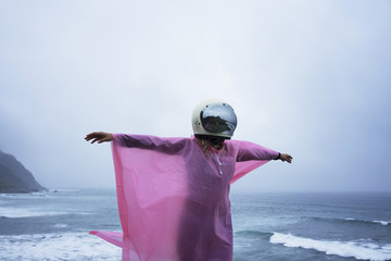 Unrecognizable girl in pink raincoat and white helmet stands above blue ocean and spreads her arms around under rain in fog day.