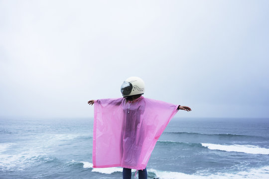 Unrecognizable girl in pink rain coat and white helmet stands above blue ocean and spreads her arms around under rain in fog day.