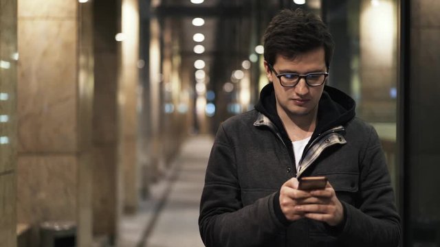 Front view of a handsome young man wearing glasses and a jacket. He is texting to somebody and smiling when he gets a reply. Locked down real time medium shot