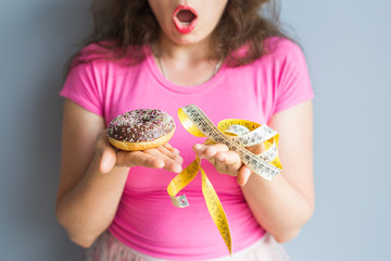 Naklejka premium Close-up of confused young woman holding a donut and a measuring tape. The concept of healthy eating. Diet.