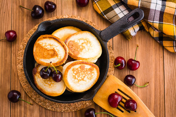 Fried pancakes in a iron pan and ripe cherries on a wooden table. Top view