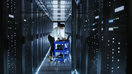 Female IT Engineer Works on a Laptop and Scans and Swaps Hard Drives That are Lying on a Cart. She Works in Data Center.