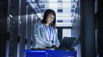 Asian Female IT Engineer Working on a Laptop on Tool Cart, She Scans Hard Drives.  She's in a Big Data Center Full of Rack Servers.