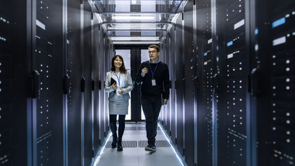 Caucasian Male and Asian Female IT Technicians Walking through Corridor of Data Center with Rows of...