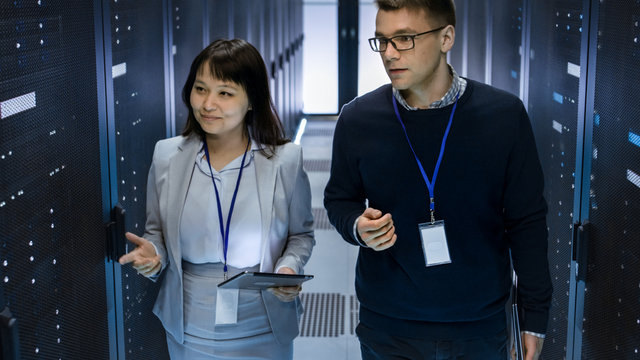 Caucasian Male and Asian Female IT Technicians Walking through Corridor of Data Center with Rows of Rack Servers. They Have Discussion, She Holds Tablet Computer.