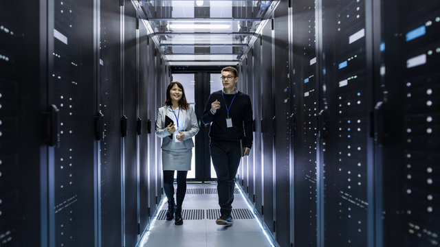 Caucasian Male and Asian Female IT Technicians Walking through Corridor of Data Center with Rows of Rack Servers. They Have Discussion, She Holds Tablet Computer.