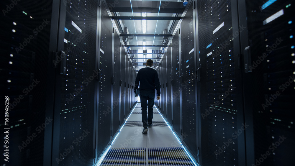 Poster following shot of it engineer walking through data center corridor with rows of rack servers.