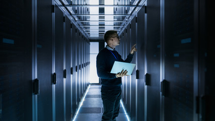 Male IT Engineer Works on a Laptop in front of Server Cabinet at a Big Data Center. Rows of Rack Servers are Seen.
