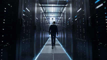 Following Shot of IT Engineer Walking Through Data Center Corridor with Rows of Rack Servers.