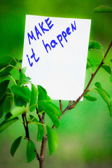 Motivating phrase make it happen. On a green background on a branch is a white paper with a motivating phrase.