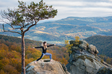 Sporty attractive female is practicing yoga and doing asana Virabhadrasana 2 on the top of the mountain near big tree. Autumn forests, rocks and hills on the background
