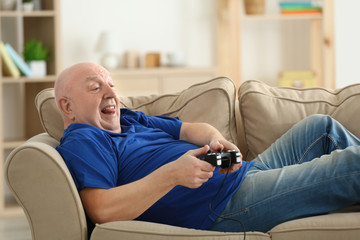 Obraz premium Fat senior man playing videogame while lying on sofa at home. Sedentary lifestyle concept