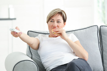 Mature woman watching TV and eating hamburger while sitting on sofa at home. Sedentary lifestyle concept