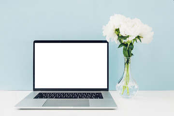 Fototapeta premium Laptop with white blank screen and flowers in vase on table on blue background. mock up