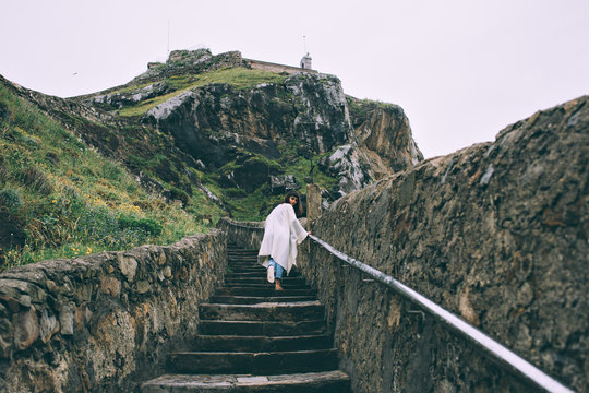 Young woman model is exploring ancient stairs on cliffs and rocks of medieval monastery or castle high in mountains, looks back and smiles at camera