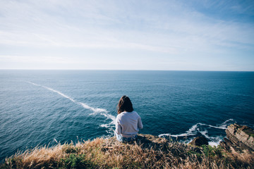 Young woman with dark hair sits on edge of cliff, dreamingly overlooks ocean waves and horizon,...
