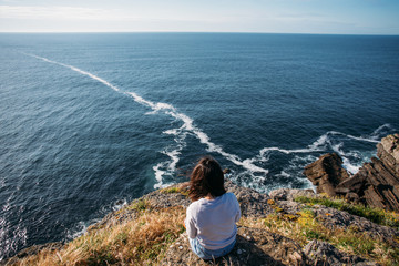 Young woman with dark hair sits on edge of cliff, dreamingly overlooks ocean waves and horizon,...