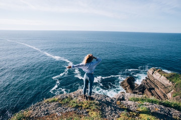 Blonde pretty woman in jeans and shirt looks at amazing ocean scenery, waves and cliffs, wind plays...